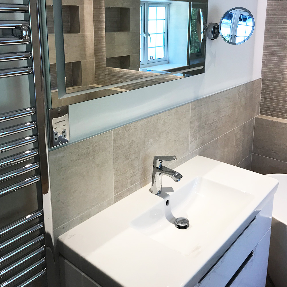 Modern bathroom with a white sink, chrome faucet, large wall mirror, small round magnifying mirror, and tiled walls. A window and towel radiator are also visible.