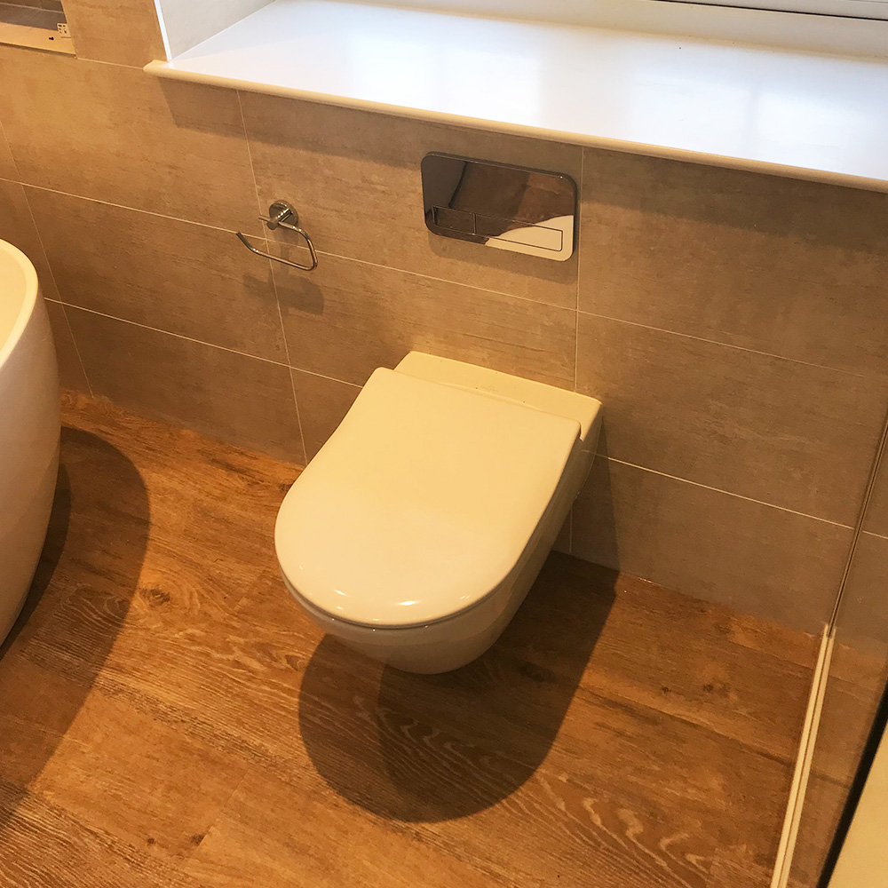 A modern wall-mounted toilet with a rectangular flush panel above it, set against beige tiled walls and a wooden floor.
