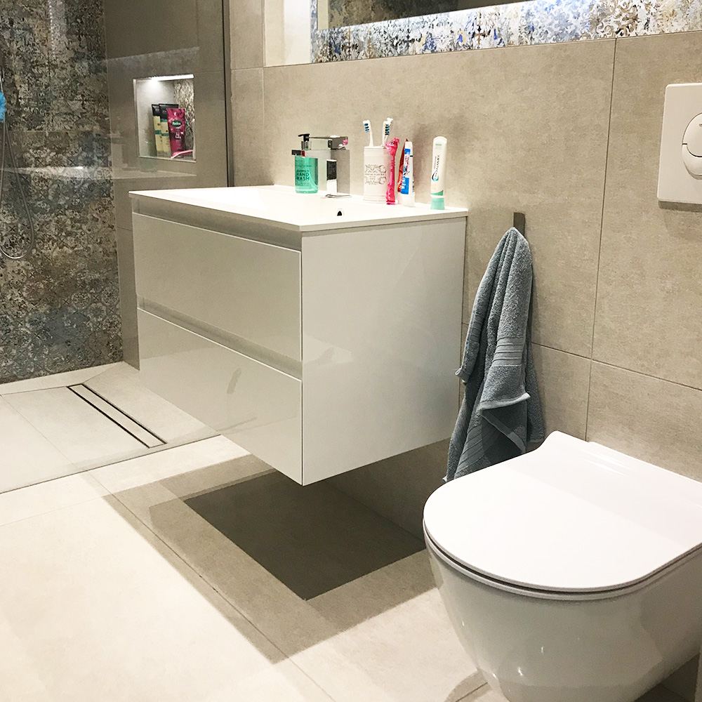 Modern bathroom with a floating white vanity, sink, toiletries, wall-mounted toilet, gray towel hanging on the side, and a glass shower enclosure in the background.