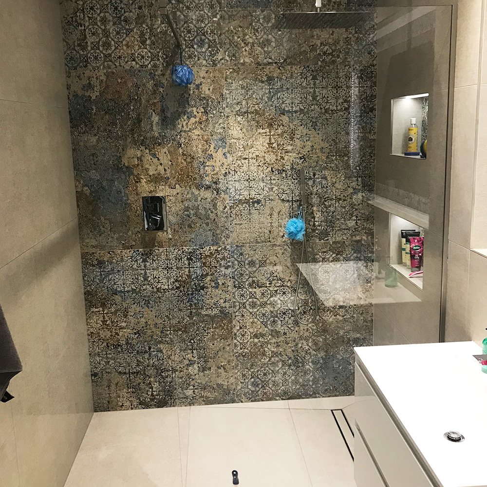 Modern bathroom shower with patterned tile wall, built-in shelf holding toiletries, glass enclosure, and white vanity visible in the foreground.