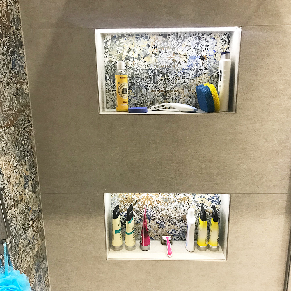 Two recessed bathroom shelves lined with toiletries, including bottles, a toothbrush, a razor, a sponge, and other hygiene products against a patterned background.