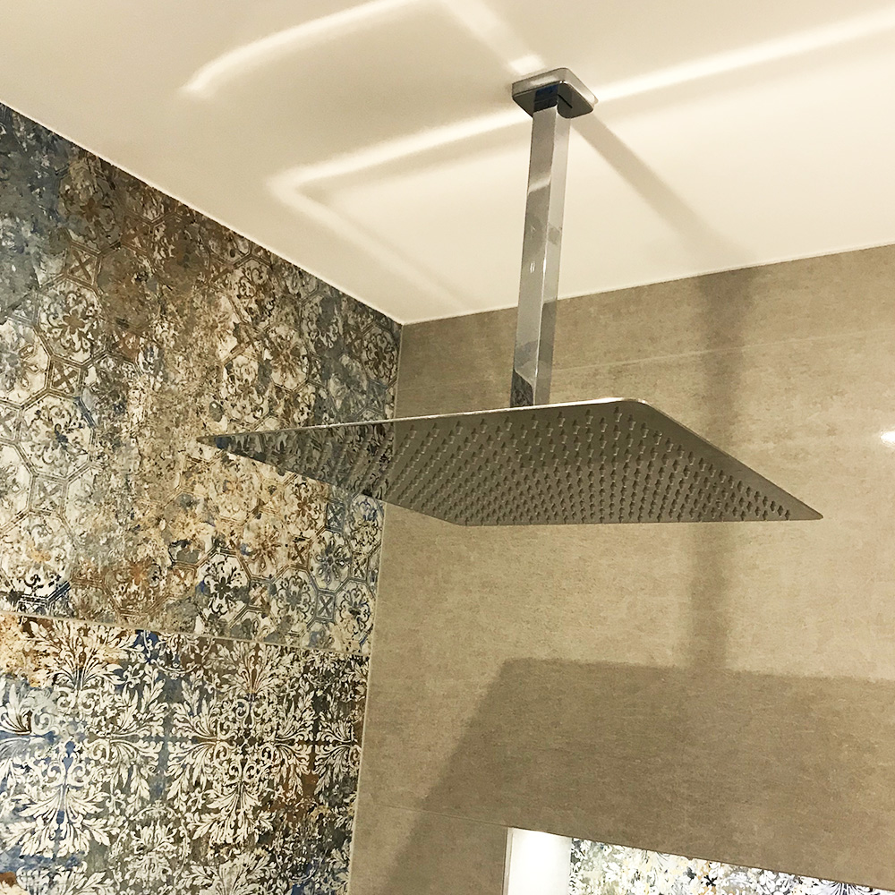 Ceiling-mounted square rainfall shower head in a bathroom with patterned and plain beige tile walls.