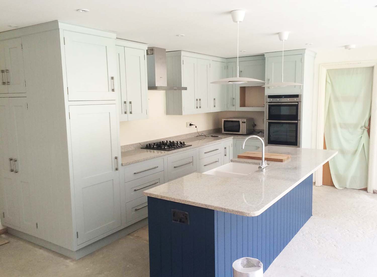 Modern kitchen with light grey cabinets, a central island with blue base, built-in oven, gas cooktop, pendant lights, and an unfinished floor.
