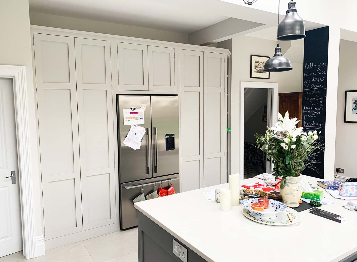 Modern kitchen with built-in cabinets, a stainless steel refrigerator, island counter with various items, vase of flowers, and pendant lights overhead.