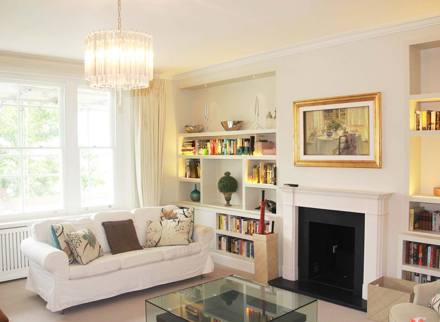A bright living room with a white sofa, glass coffee table, bookshelves, a fireplace, and a large window letting in natural light.