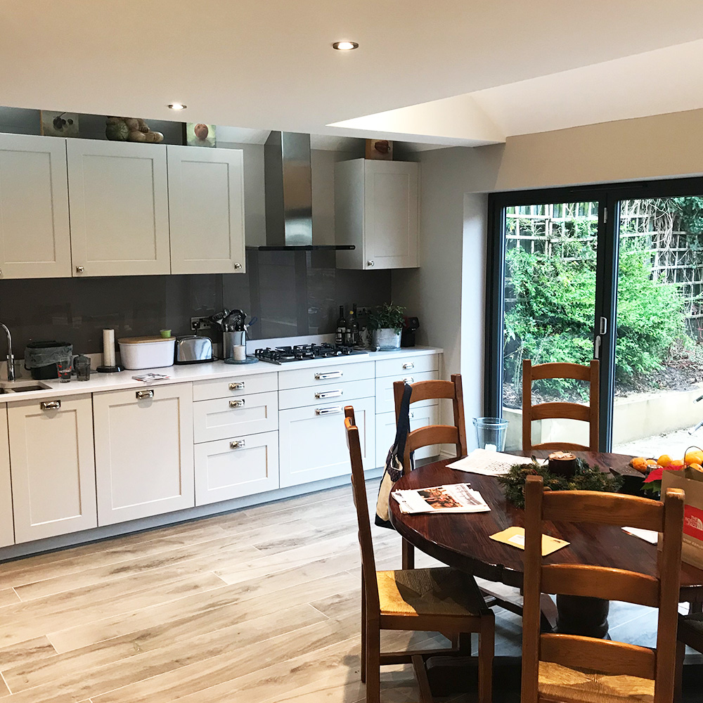 Modern kitchen with white cabinets, stainless steel appliances, and a round wooden dining table with four chairs, set near large glass doors overlooking a garden.
