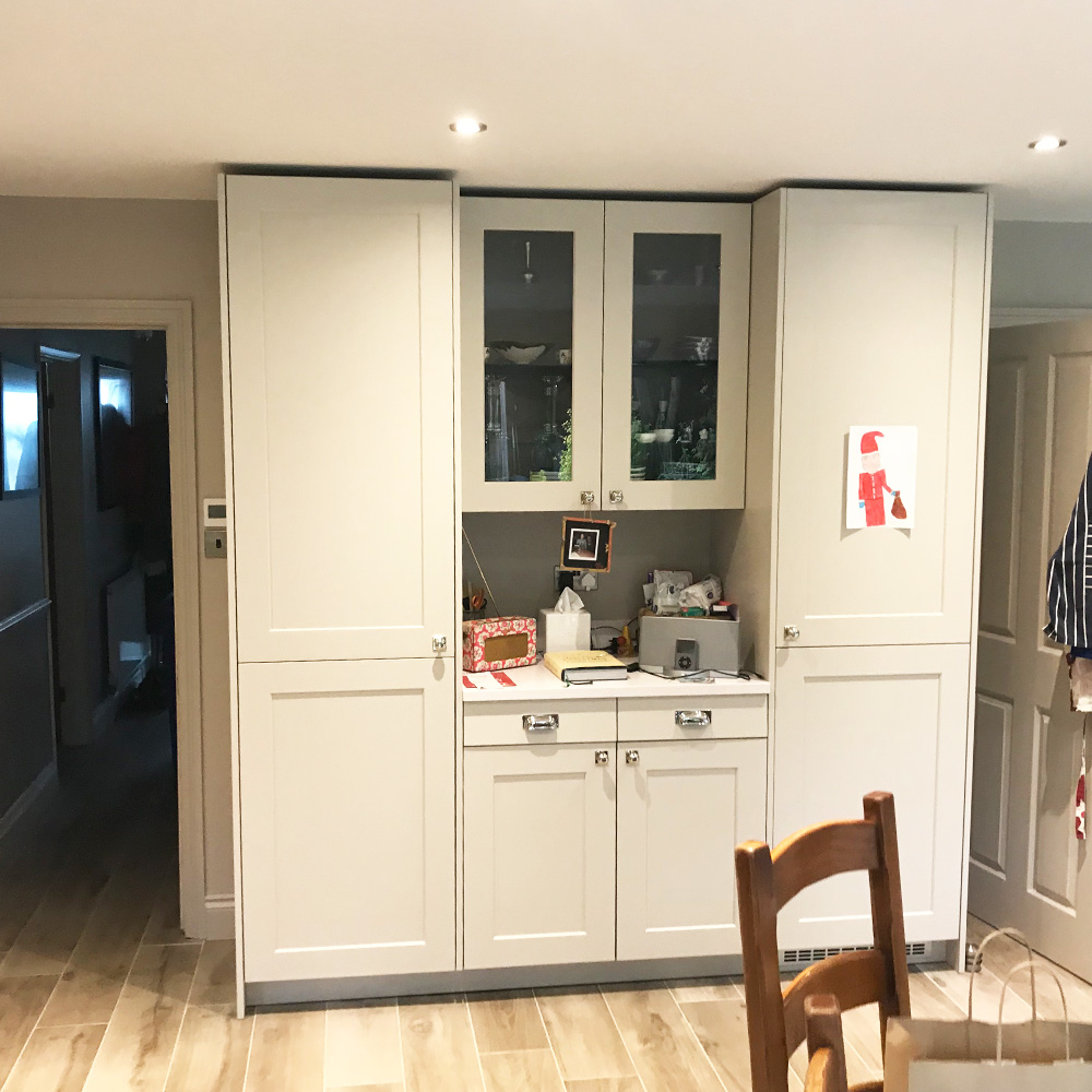 A modern kitchen unit with light-colored cabinets, glass-fronted upper doors, and various items on the countertop and shelves. A child's drawing is attached to the right side cabinet.
