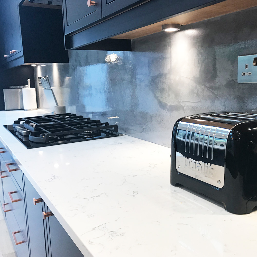A modern kitchen countertop with a black toaster, gas stove, white marble surface, and metallic backsplash under cabinet lighting.