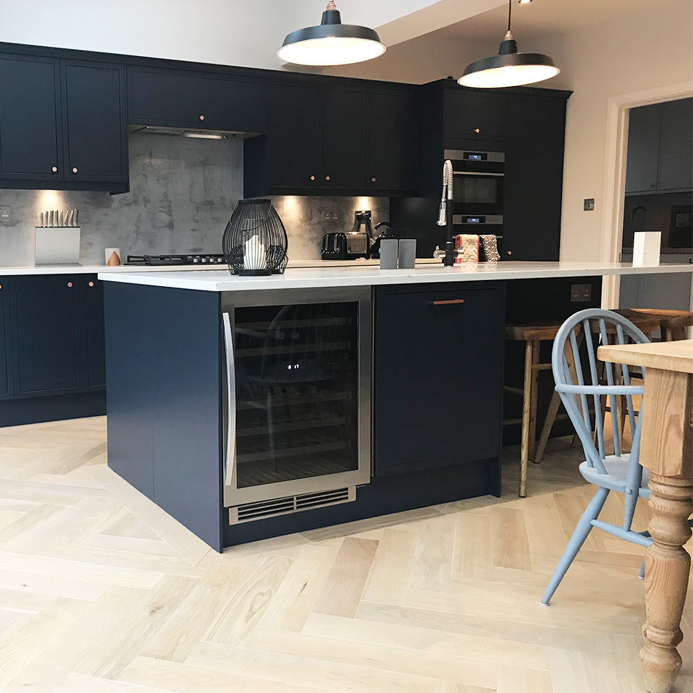 Modern kitchen with dark blue cabinets, light wood herringbone floor, a central island with a wine cooler, pendant lights, and a small dining area with blue chairs.