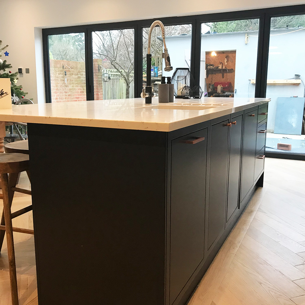 A modern kitchen island with a marble countertop, dark cabinetry, sink, and faucet, situated near large glass doors overlooking a backyard.