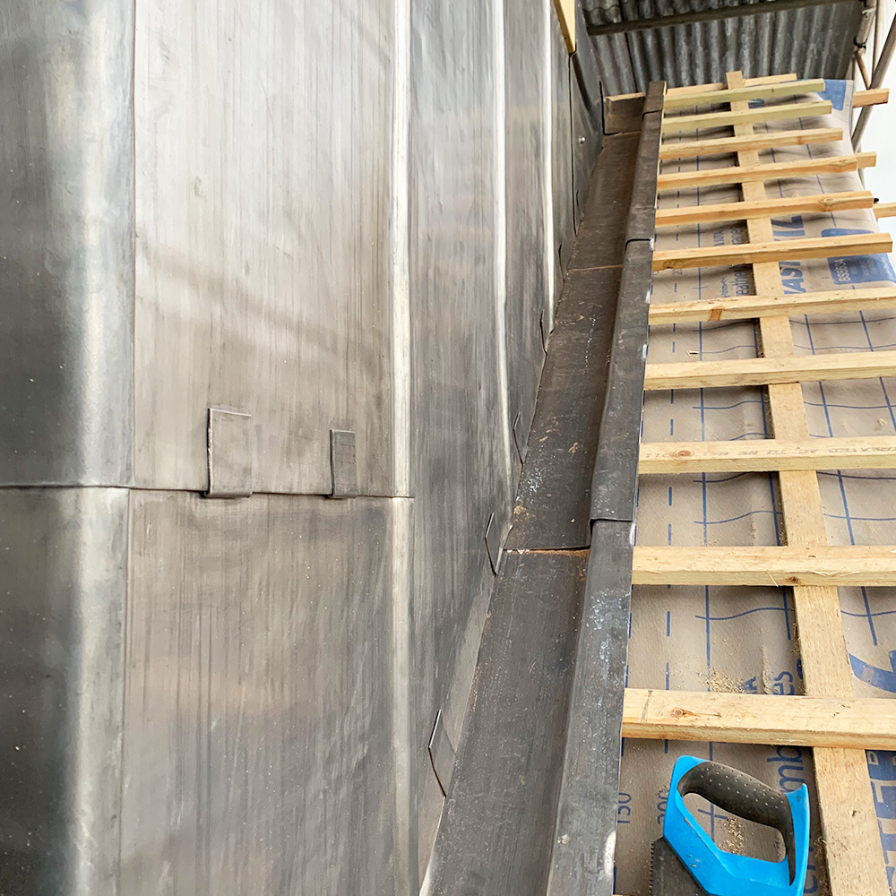Close-up of a metal roof with overlapping panels, a gutter channel, and a wooden framework under construction. A blue utility knife is on the right.