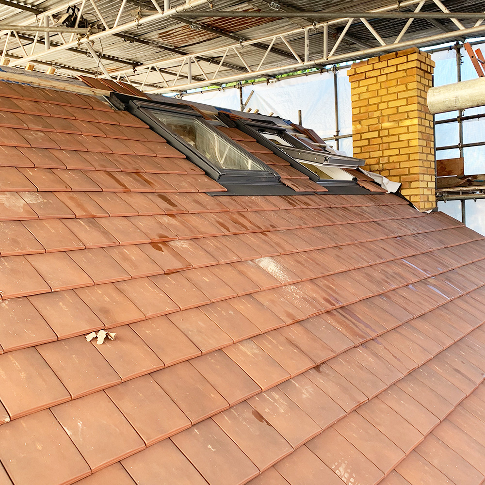 A sloped roof with reddish-brown tiles, a skylight window, and a yellow brick chimney under scaffolding.