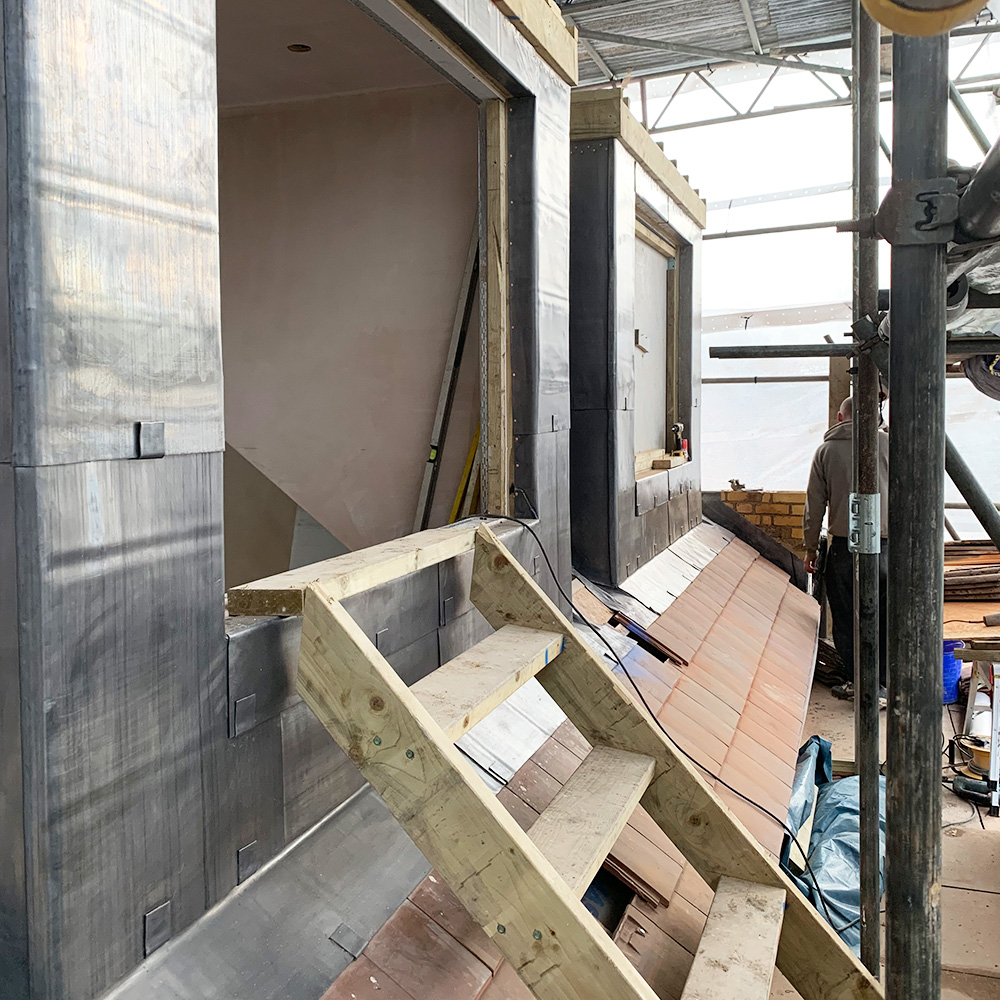 Interior view of a building under construction with wooden stairs, unfinished walls, exposed insulation, and construction materials scattered around.